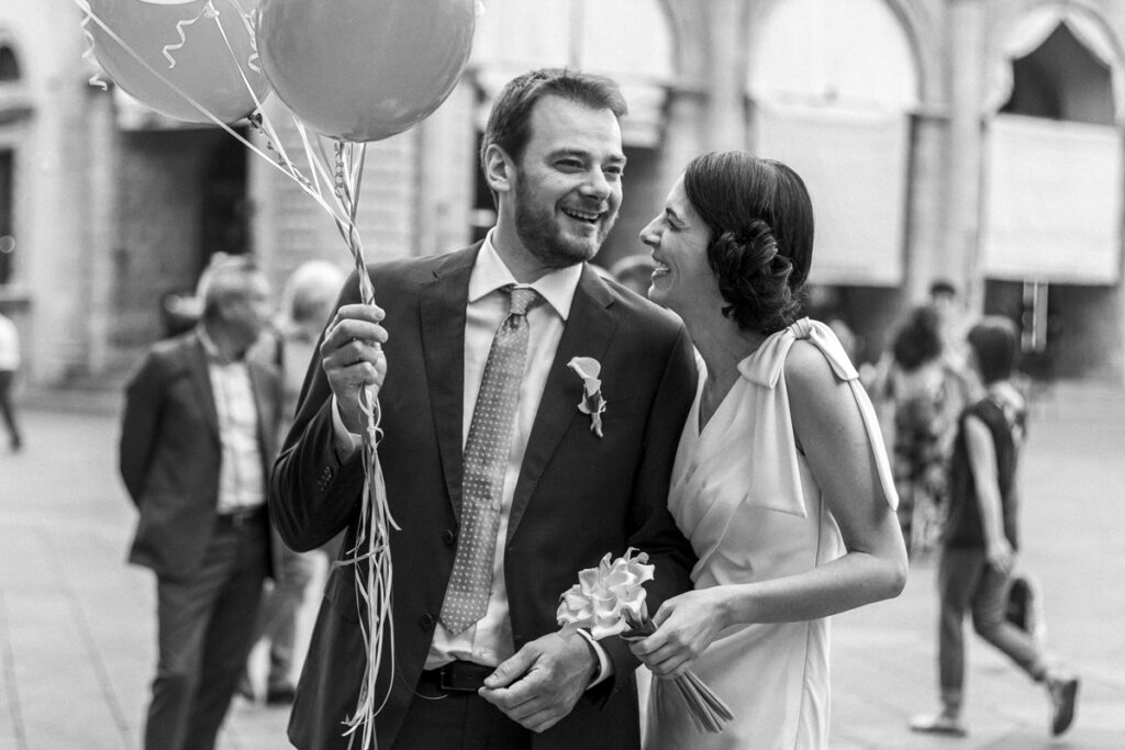 the newlyweds walk in piazza Maggiore in Bologna smiling and with balloons